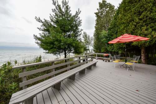 A wooden deck by the water with chairs, an umbrella, and trees, overlooking a calm lake under a cloudy sky.