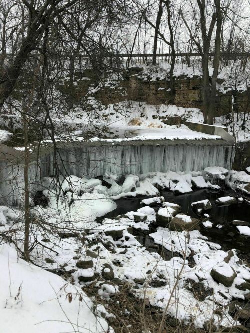A frozen waterfall surrounded by snow-covered rocks and trees in a winter landscape.