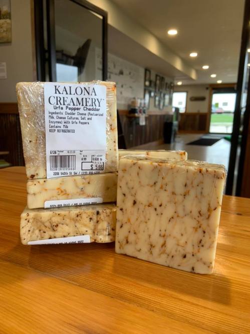 A stack of Kalona Creamery Urfa Pepper Cheddar cheese blocks on a wooden table, with packaging visible.