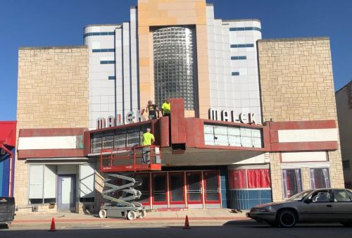 Workers on a lift are renovating the marquee of the Malick Theater, a vintage building with art deco architecture.
