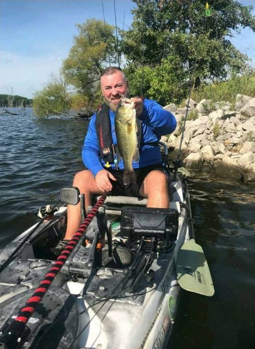 A man in a kayak holds up a large fish, smiling, with a scenic lake and trees in the background.
