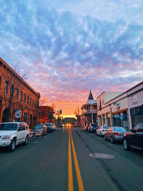 A vibrant sunset paints the sky over a charming street lined with shops and parked cars.