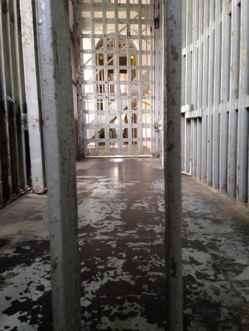 View through prison bars into a dimly lit corridor with metal grates and a staircase in the background.