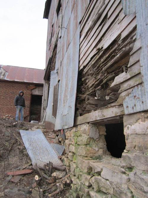 A person stands near a dilapidated barn with peeling metal and crumbling stone walls, surrounded by debris.