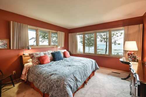 Cozy bedroom with a large bed, warm orange walls, and windows overlooking a scenic view of the water and trees.