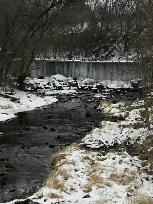 A snowy landscape with a frozen waterfall, a stream flowing through rocks, and bare trees in the background.