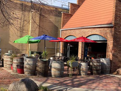 Outdoor seating area with colorful umbrellas, surrounded by wooden barrels, next to a brick building.