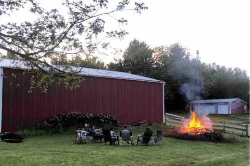 A group of people sitting around a large bonfire near a red barn at dusk, with trees in the background.