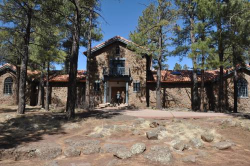 A large stone house with a red tile roof, surrounded by tall trees and rocky ground under a clear blue sky.