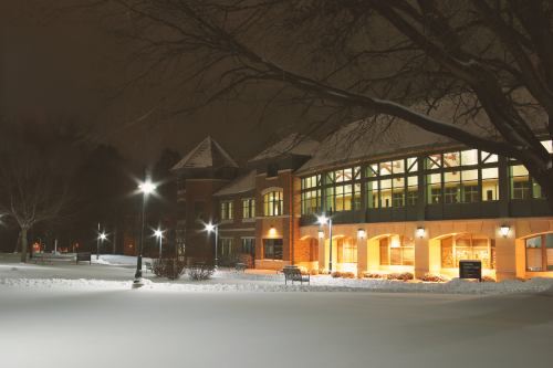 A snow-covered building at night, illuminated by warm lights, surrounded by trees and a quiet, snowy landscape.