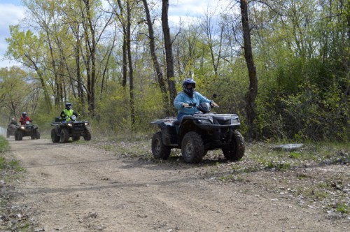 Three people riding all-terrain vehicles on a dirt path surrounded by trees and greenery.
