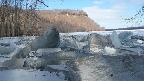 Ice chunks scattered along a frozen riverbank, with a wooded hillside in the background under a partly cloudy sky.