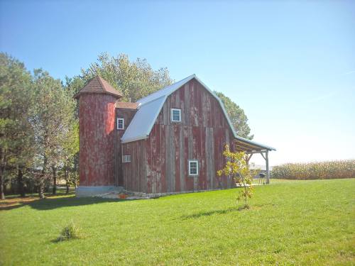 A red barn with a silo, surrounded by green grass and trees, under a clear blue sky.