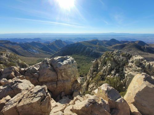 A panoramic view of rugged mountains under a bright sun, with rocky foreground and distant peaks in the background.