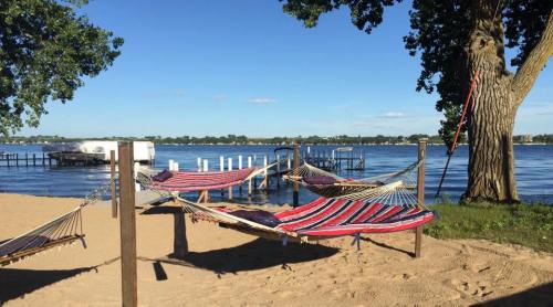 Three colorful hammocks are set up on a sandy beach by a calm lake, with a dock and trees in the background.