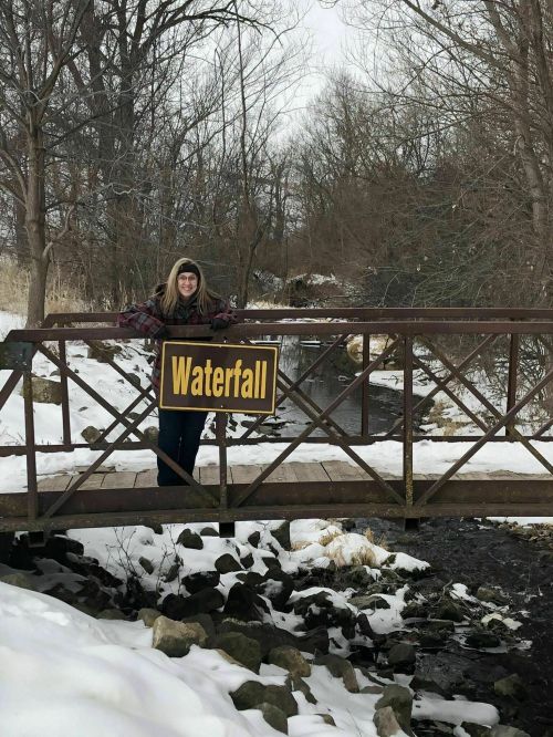 A person stands on a bridge by a snowy creek, holding a sign that reads "Waterfall." Trees are bare in the background.