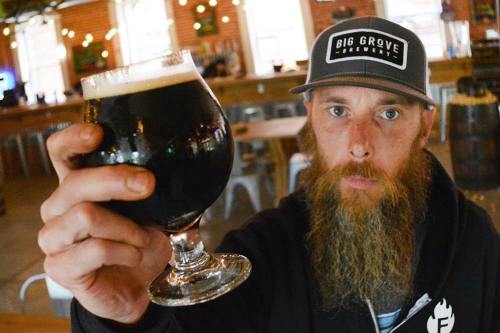 A bearded man in a cap holds up a dark beer in a glass, inside a brewery with wooden decor.