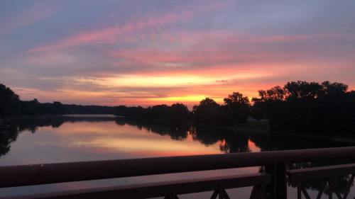 A serene sunset over a calm river, with colorful clouds reflecting on the water and silhouettes of trees in the background.