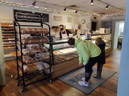 A bakery interior with a display of fresh bread. A customer in a green hoodie examines the goods while a staff member assists.