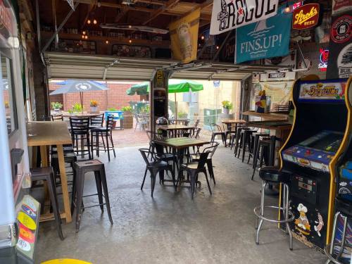 A rustic bar interior with tables, chairs, and an arcade game, featuring garage doors and colorful banners.