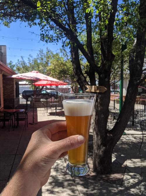 A hand holding a tall glass of beer in a sunny outdoor setting with trees and patio furniture in the background.