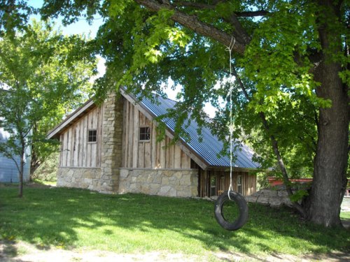 A rustic wooden cabin with a stone foundation, surrounded by trees and a tire swing hanging from a branch.