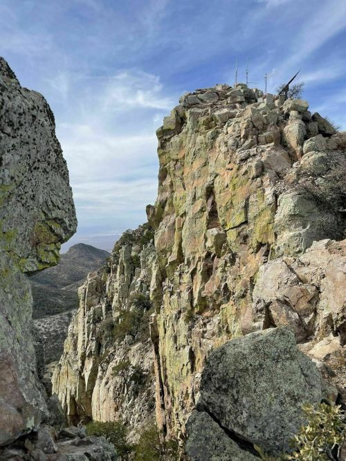 A rugged rock formation with antennas on top, surrounded by hills and a blue sky with wispy clouds.