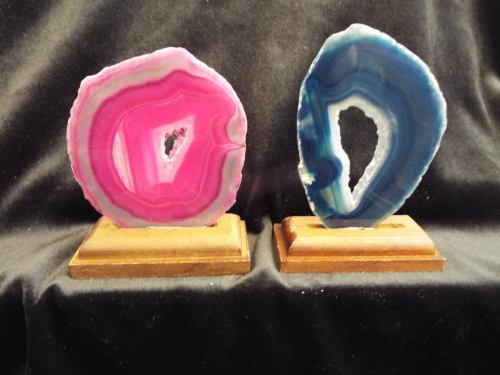 Two polished agate slices, one pink and one blue, displayed on wooden stands against a black background.