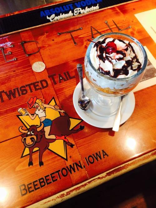 A dessert in a glass topped with whipped cream and a cherry, placed on a wooden bar with a cowboy-themed logo.