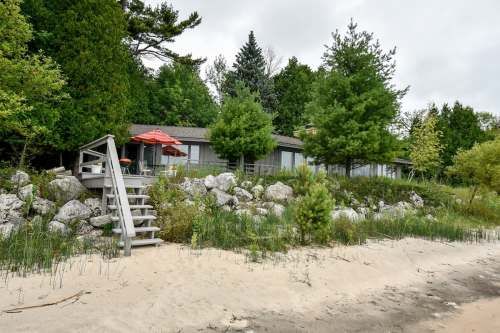 A beach house surrounded by trees, with a sandy shore and a staircase leading up to a patio with an umbrella.