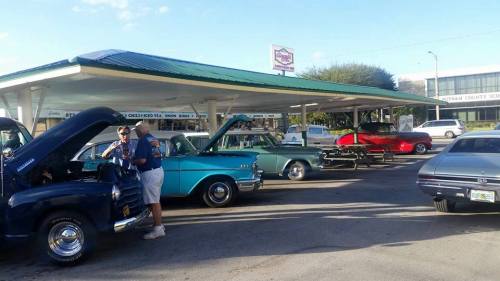 Classic cars on display at a car meet, with people chatting and working on vehicles under a shaded pavilion.