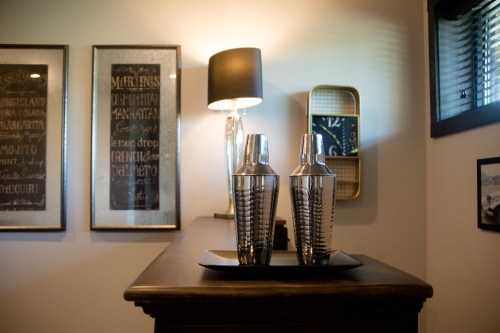 A stylish bar setup featuring two cocktail shakers on a tray, with framed drink menus and a lamp in the background.