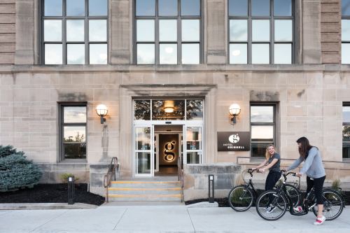 Two women stand with bicycles outside the entrance of Hotel Grinnell, featuring large windows and a welcoming facade.