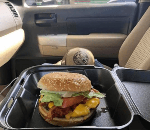 A close-up of a cheeseburger with bacon, lettuce, and tomato in a takeout container, set inside a car.