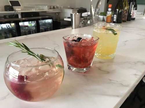 Three colorful cocktails on a marble bar top: a pink drink with rosemary, a red drink with berries, and a yellow drink with mint.