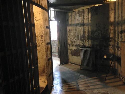 A dimly lit, empty jail cell with a barred door, peeling walls, and a metal chair in the corner.