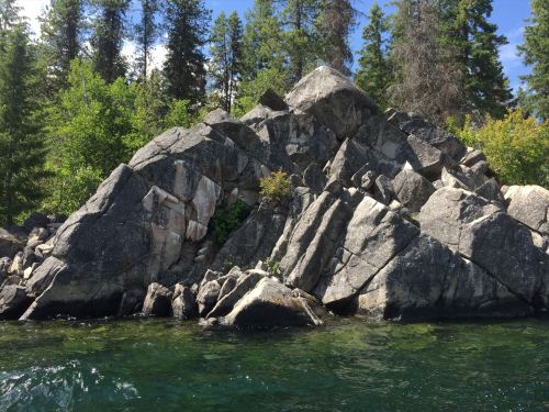 A rocky shoreline with large boulders and green trees under a clear blue sky. Water reflects the landscape.