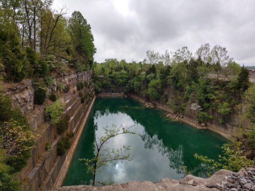 A serene, green-tinted quarry pond surrounded by rocky cliffs and lush trees under a cloudy sky.