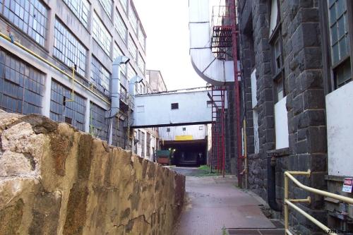 A narrow passage between industrial buildings, featuring stone walls and large windows, with a glimpse of a truck in the distance.