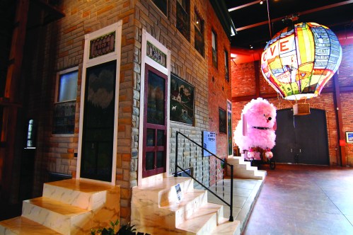 A whimsical indoor scene featuring a colorful hot air balloon and a large fluffy pink sculpture beside a brick building.