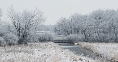 A serene winter landscape featuring a frosty river, a wooden bridge, and snow-covered trees.