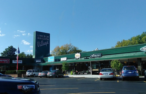 A shopping plaza with a sign for Lincoln Heights Village, featuring various storefronts and a clear blue sky.