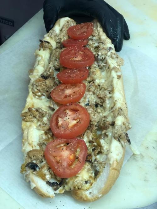 A large sub sandwich topped with sliced tomatoes, chicken, cheese, and other toppings, held by a gloved hand.