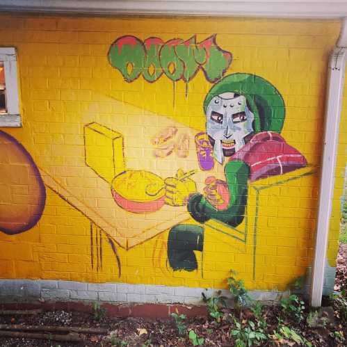 A colorful mural of a masked figure sitting at a table with food and drinks, set against a bright yellow wall.