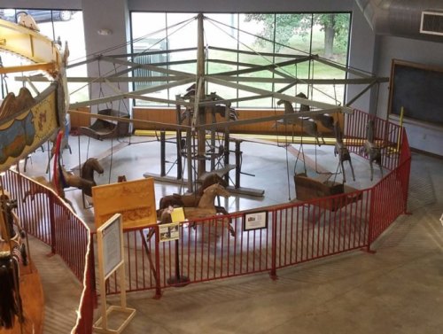 An empty carousel with wooden horses, surrounded by a red fence, inside a spacious room with large windows.