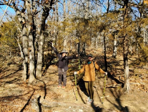 Two hikers on a wooded trail, one raising arms in excitement, both holding trekking poles, surrounded by trees.