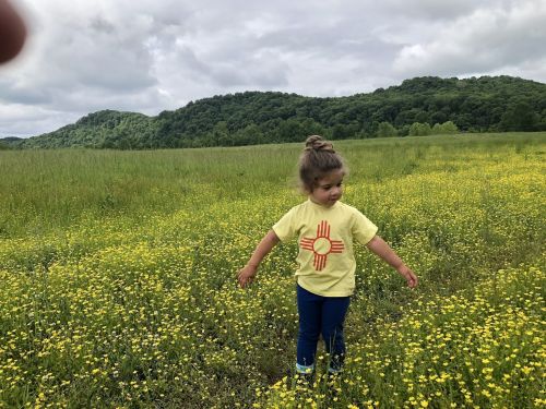 A young child in a yellow shirt with a design stands in a field of yellow flowers, surrounded by green hills.