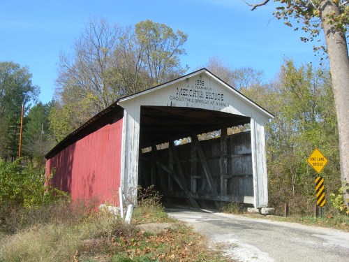 A red covered bridge with a wooden interior, surrounded by trees and a sign indicating it's a one-lane bridge.