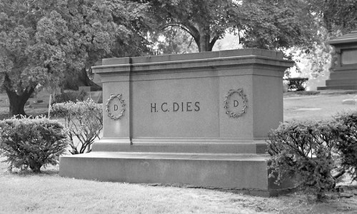 A gray tombstone in a cemetery, engraved with "H.C. DIES," surrounded by trees and shrubs.