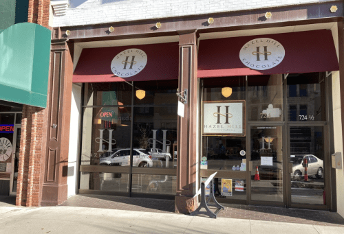 Exterior of Hazel Hill Chocolate shop, featuring large windows, a red awning, and an "Open" sign.